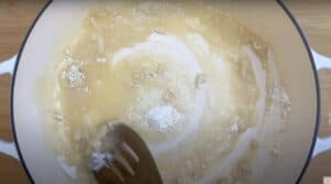 Stirring flour and butter to make a smooth roux for sauce in a white pan on a wooden surface.